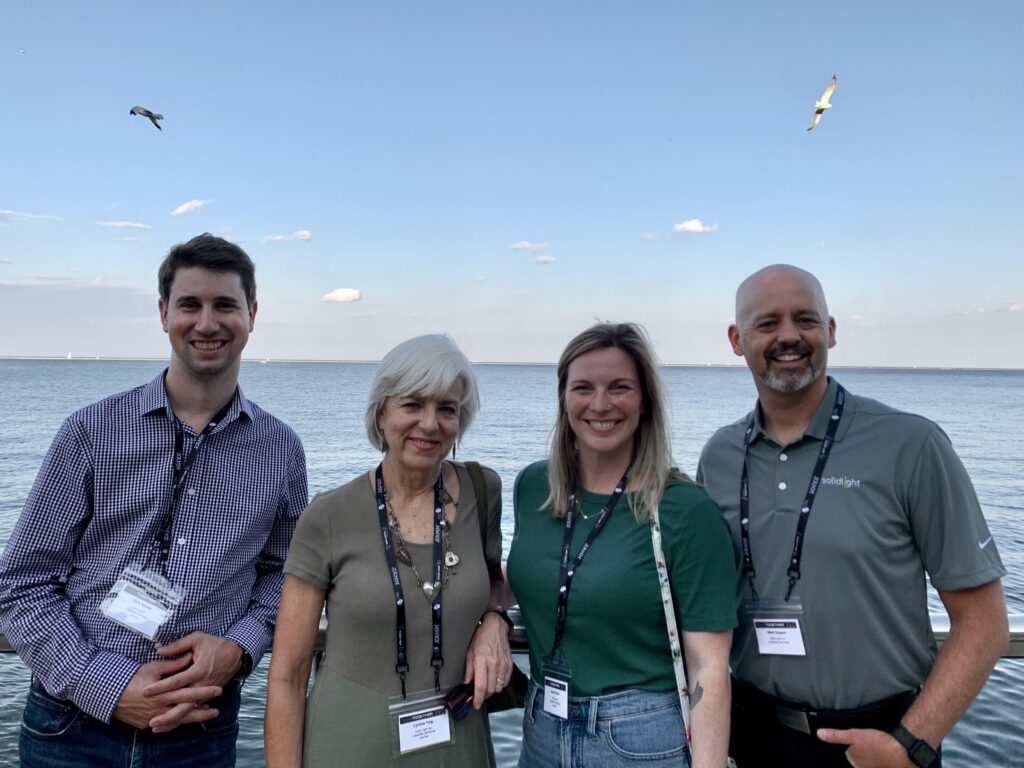Photo depicts Solid Light team members, Chris Mozier, Cynthia Torp, Jamie Glavic, and Mark Sargent, wearing their conference badges against a backdrop of Lake Michigan in Milwaukee.